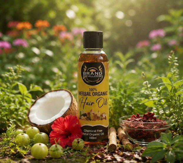 A bottle of Brand of Outfits Herbal Organic Hair Oil stands outdoors on a grassy ground surrounded by green foliage, a cut coconut, green berries, a red flower, and a small bowl of dried ingredients.

